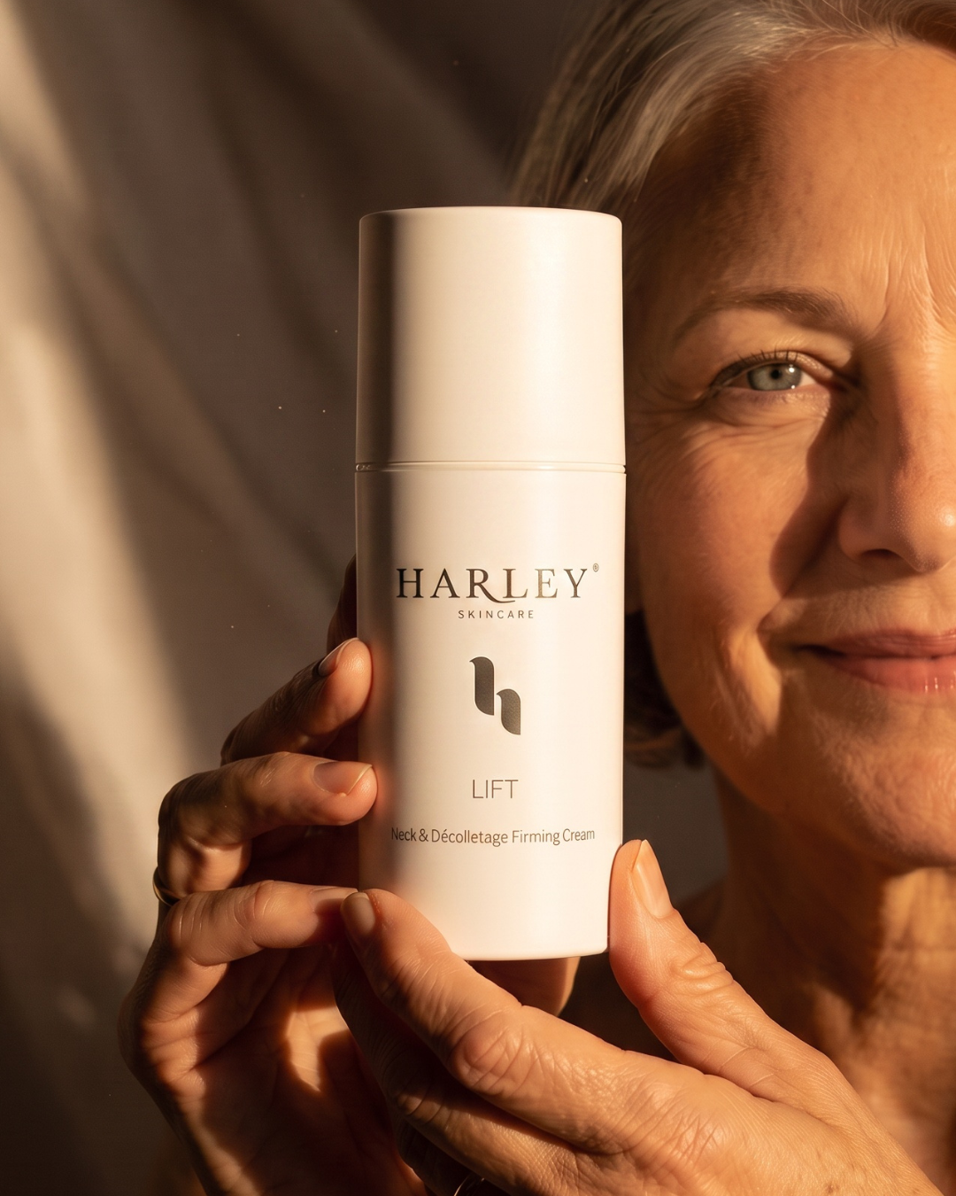 A woman holding a white cylindrical bottle of Harley Skincare Lift Neck & Decolletage Firming Cream, with warm sunlight casting shadows on her face and the bottle.