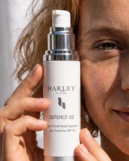 A woman with wavy light brown hair holds a bottle of Harley Defence 50 Daily Tinted SPF 50 in front of her face against a white curtain background.