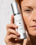 A woman with short red hair holds a bottle of Harley Skincare Daily Moisturizer Sun SPF 20 in front of her face against a white textured background.