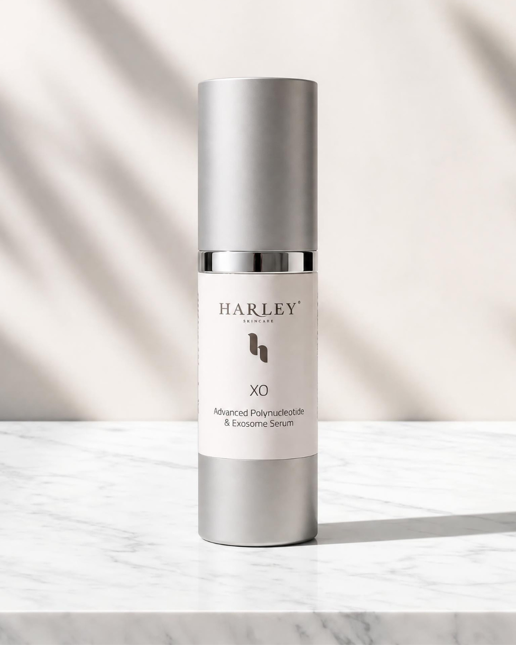 A Harley Skincare X0 Advanced Polynucleotide & Exosome Serum bottle, featuring a sleek silver metallic design with a white label, set on a white marble surface with soft leaf shadows.