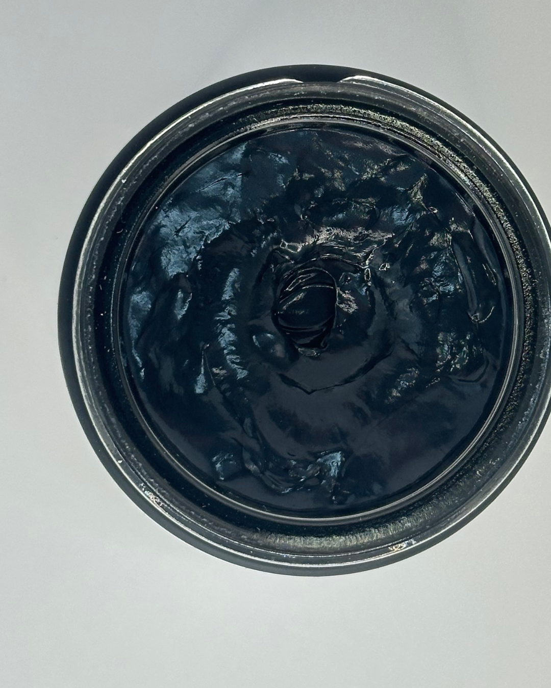 A close-up top-down view of a dark, glossy, thick cosmetic cream or balm inside a round black container, set against a plain light gray background.