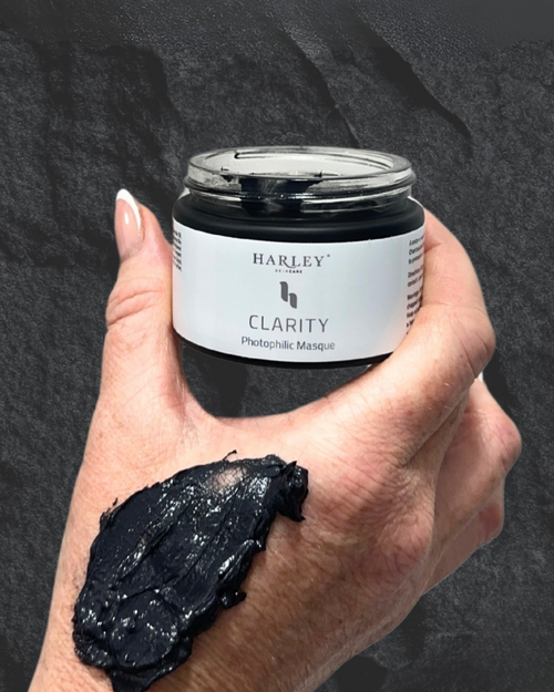 A hand holding a small jar of Harley Skincare Clarity Charcoal Mask, with a dollop of the black mask applied to the back of the hand, set against a dark textured background.