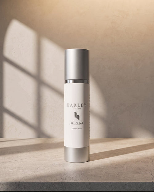 A cylindrical Harley Skincare All-Clear Hydrating Mask bottle with a white body and metallic silver cap, standing on a stone surface with warm sunlight and window shadow patterns on a textured beige wall.