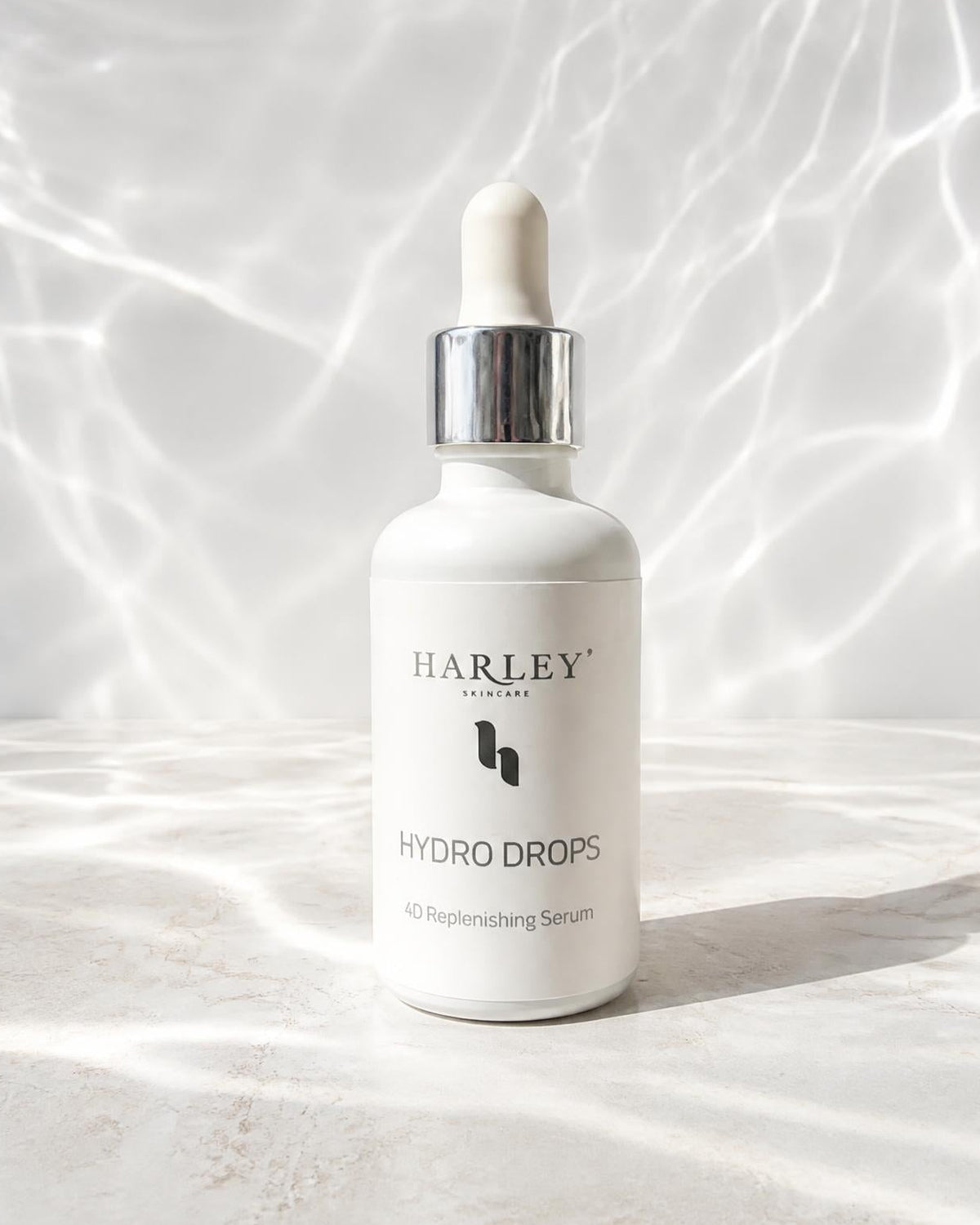 A white dropper bottle of Harley Skincare Hydro Drops serum with a silver collar and white dropper cap, set on a light marble surface with dappled light patterns on a white background.