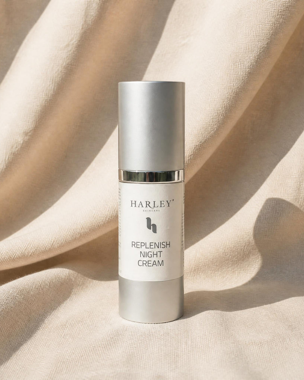 A silver cosmetic bottle labeled 'HARLEY skincare REPLENISH NIGHT CREAM' standing upright on draped beige fabric with soft shadows.