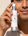 A man holding a white bottle of Harley Skincare Sensi-Clear quickly absorbing facial cleanser against a light beige textured wall.