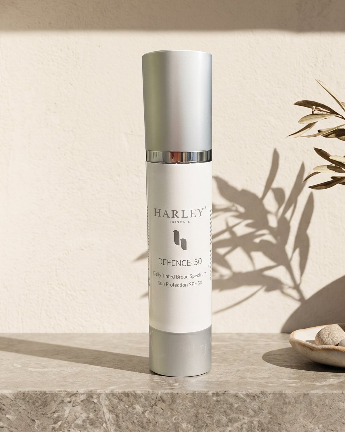 Harley Defence SPF 50 sunscreen in a silver and white cylindrical bottle, displayed on a stone shelf next to a ceramic vase with dried leaves.